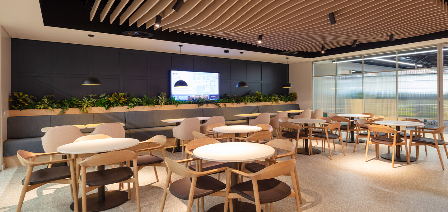 A modern well-lit cafeteria featuring round and rectangular wooden tables with matching chairs arranged neatly. A large digital screen displaying a presentation is mounted on a dark wall with a green plant shelf beneath, and large windows on one side allow natural light to fill the space.