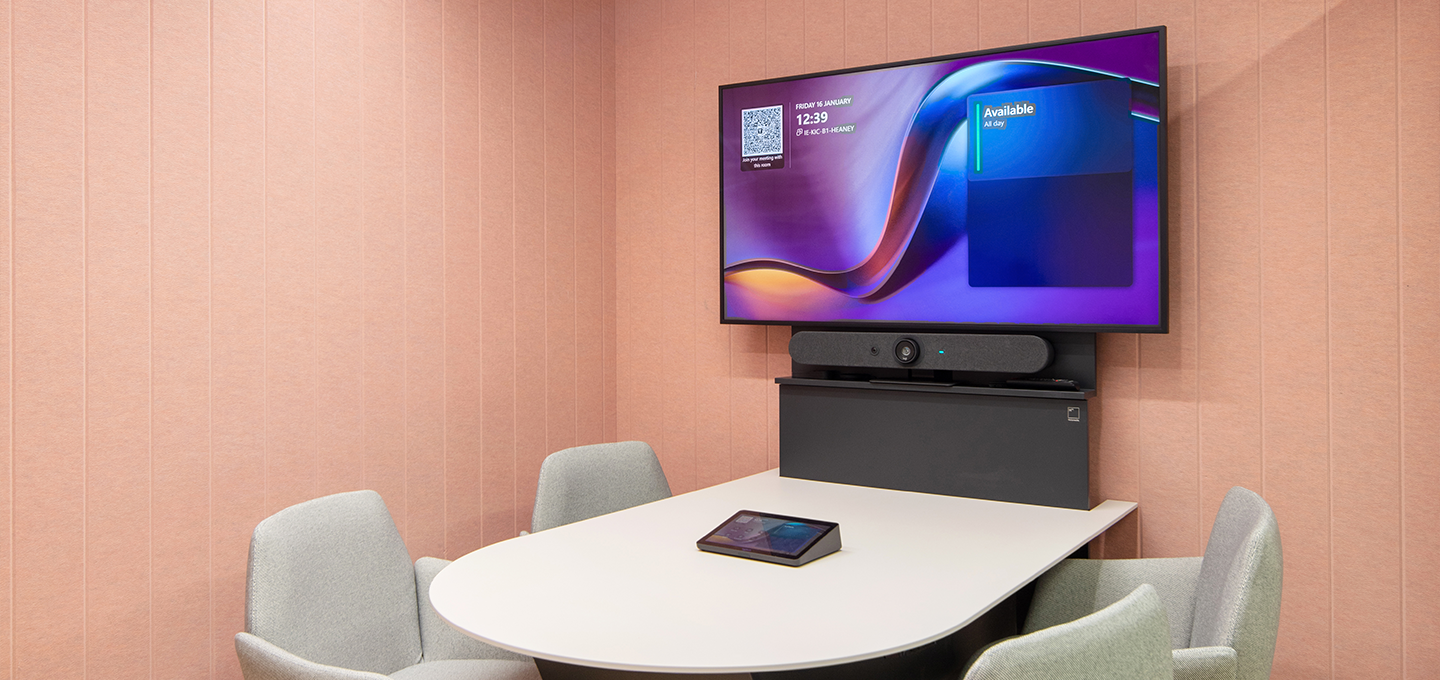 Photograph of a small conference room featuring a white oval table surrounded by four gray upholstered chairs. A large flat-screen monitor mounted on a black stand displays a purple and blue interface with a QR code, time, and availability status, while a tablet rests on the table, indicating a modern meeting or collaboration setup.