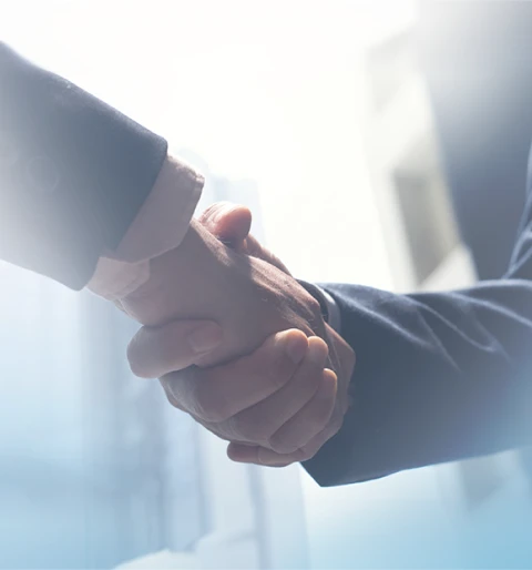 Close-up of two business professionals shaking hands, wearing suits, with a blurred modern office background.