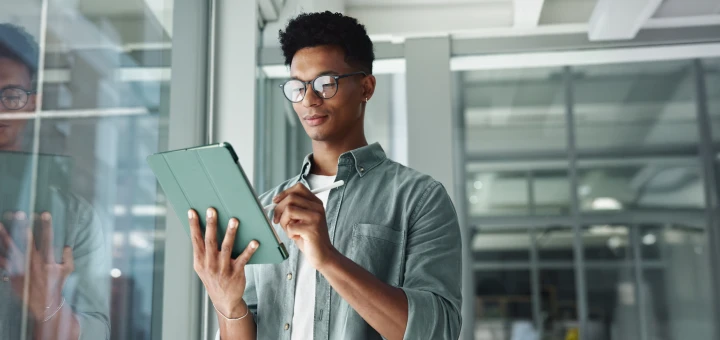 A person with glasses uses a stylus on a tablet in a modern office. They appear focused and thoughtful, surrounded by glass walls and soft lighting.