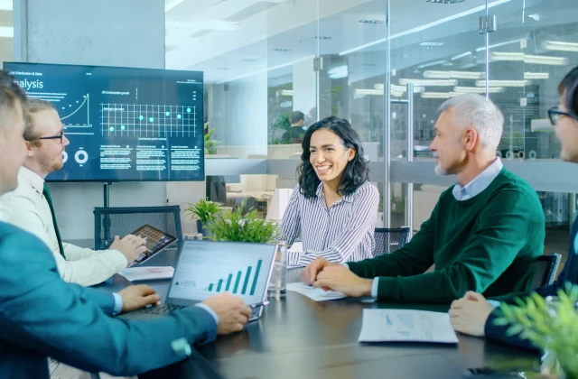  A diverse group of five business professionals are seated around a conference table in a modern office, looking at a large display screen showing financial graphs and data. A woman in the center smiles.